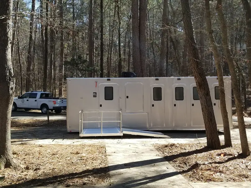 A large white ADA restroom trailer with a ramp is set up in a wooded area; a white pickup truck is parked nearby.