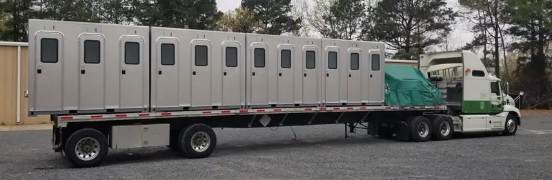 A semi-truck and flatbed trailer carrying multiple ADA restroom units, parked on a gravel lot with trees and a building in the background.