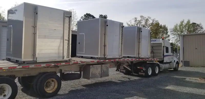 A flatbed truck loaded with three large, silver, metal restroom trailer enclosures is parked on a gravel lot next to a beige industrial building.