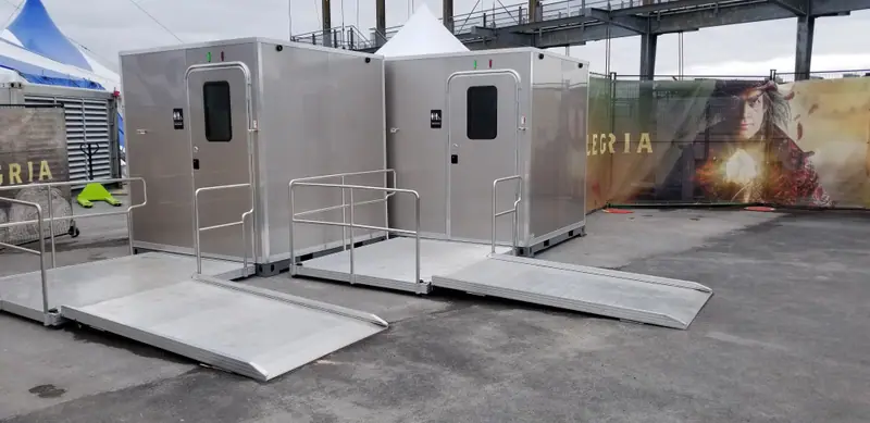 Two ADA restroom units with ramps for wheelchair access, positioned on a paved outdoor area near a decorative fence and tents.