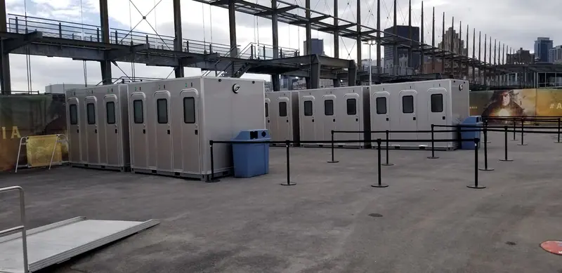 A row of portable restroom units, including an ADA restroom, stands with a blue trash bin and black queue barriers in an outdoor urban area.