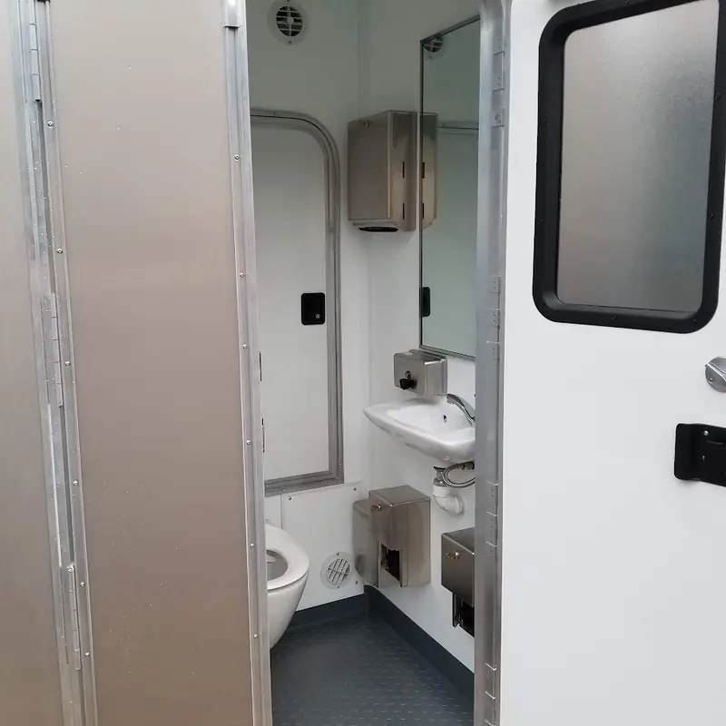 A compact ADA restroom with a toilet, sink, mirror, and mounted dispensers, viewed from the doorway. The interior of this restroom trailer features grey flooring and white walls.