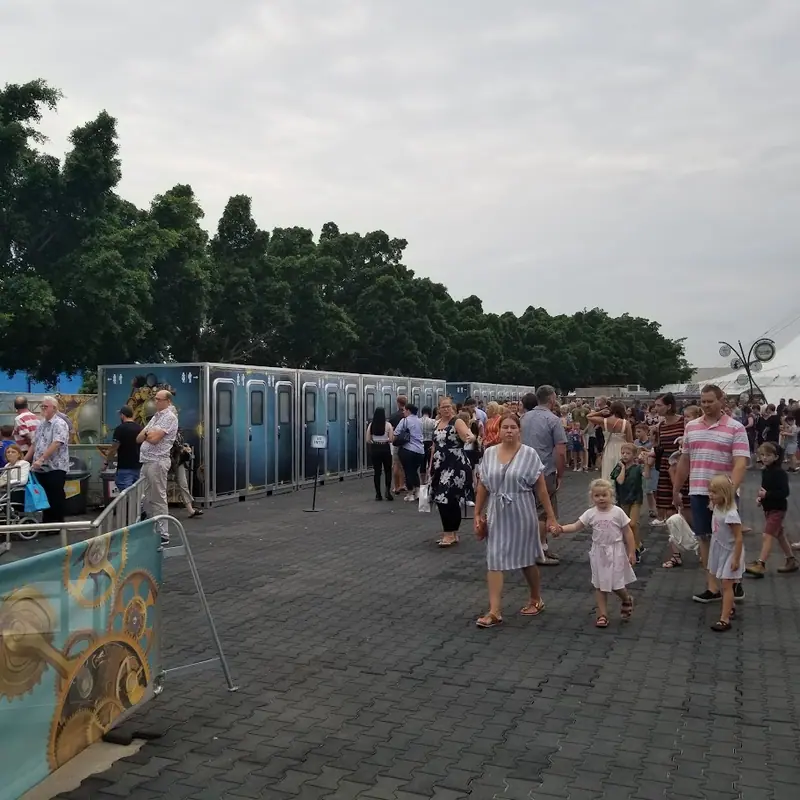 A crowd of people, including families with children, walk on a paved path near a row of portable toilets and an ADA restroom trailer on a cloudy day.