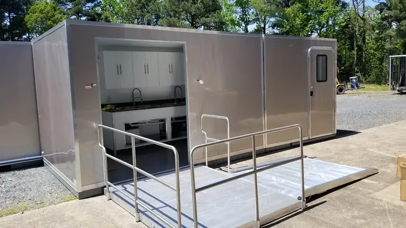 A portable modular building with a metal ramp leads to an open door, revealing an interior with white cabinets and sinks. This ADA restroom trailer sits near trees and construction equipment outside.