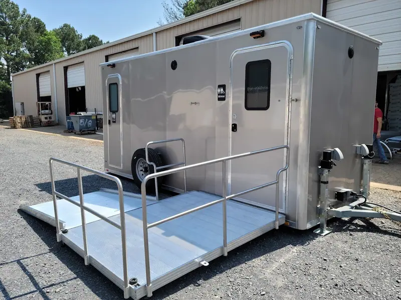 ADA restroom trailer with a metal ramp, parked outside a beige industrial building on a gravel lot.