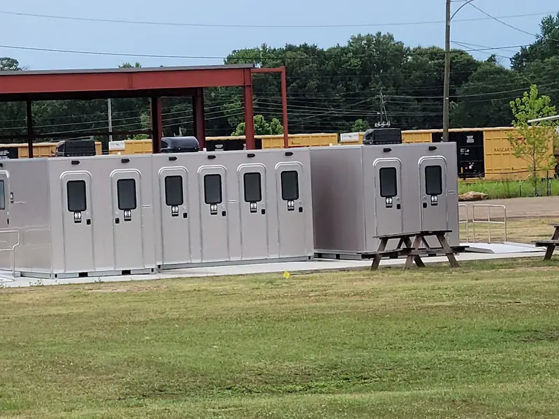 A row of portable restroom units, including an ADA restroom, is set up on a concrete pad near picnic tables, with train cars and trees visible in the background.