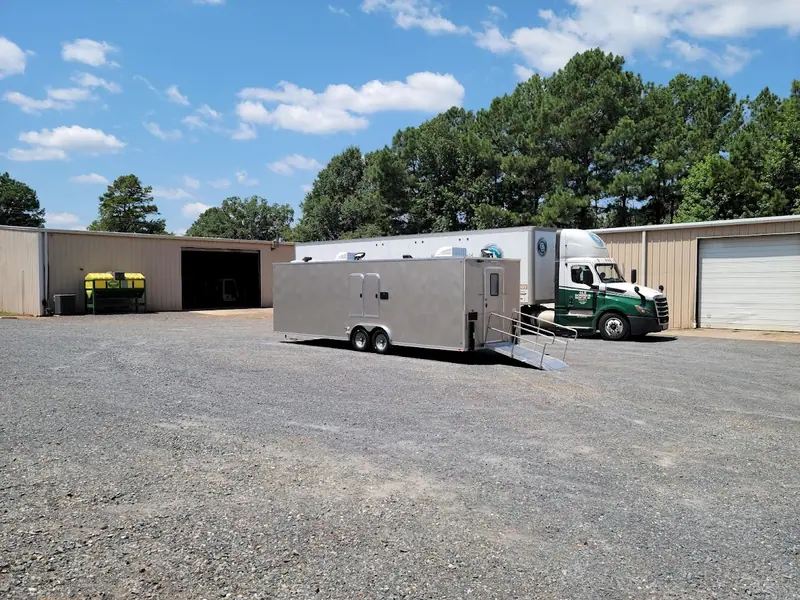 A silver ADA restroom trailer with a ramp is parked on a gravel lot near beige industrial buildings, a green semi-truck, and trees under a partly cloudy sky.