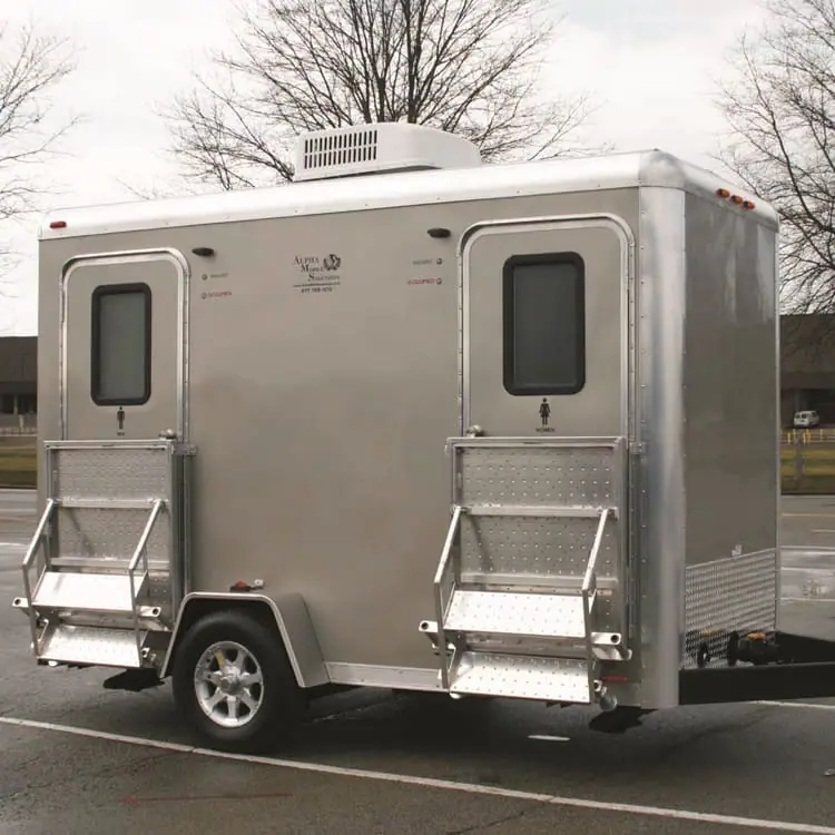 An Alpha II Restroom Trailer - A12RT with two doors and metal steps is parked in a lot with bare trees and buildings in the background.