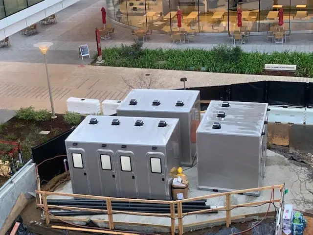 A CI 12 - 4 Stall Restroom Skid (12 ft) is set up on a construction site, surrounded by fencing, pipes, and landscaping.