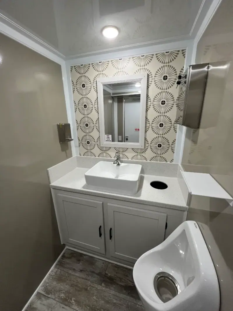 Clean ada restroom trailer featuring a sink, mirror, patterned wall, urinal, paper towel dispenser, and white cabinetry on a wood-patterned floor.