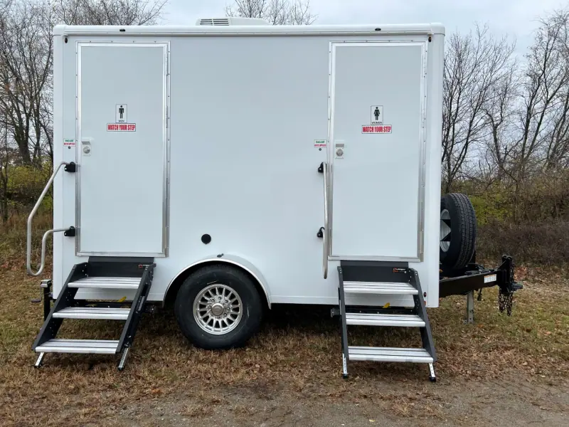 The OI-212 — 2 Stall Restroom Trailer (12' x 6') is a white unit with metal steps leading to two clearly labeled ADA restroom entrances, parked on grass.