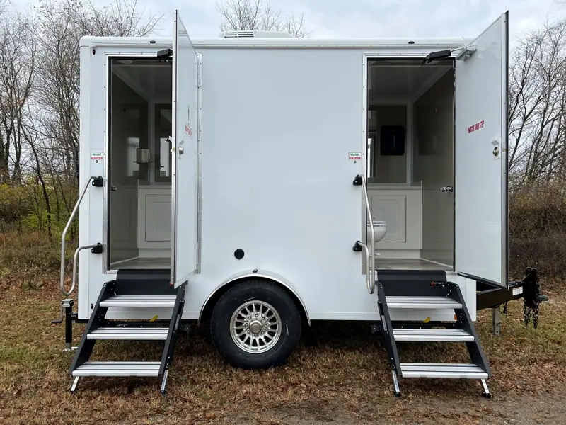 The OI-212 — 2 Stall Restroom Trailer (12' x 6') is shown with both doors open and steps to separate stalls, set on grassy ground with trees behind.