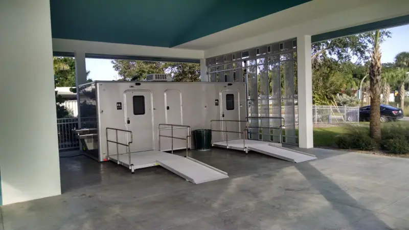 Two portable restroom trailers, including an ADA restroom trailer with attached ramps, are positioned under a covered pavilion in an outdoor setting.