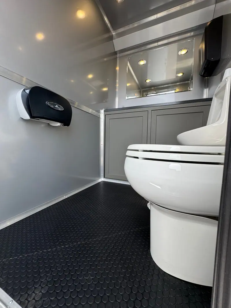 The 3 private room restroom trailer 16′ x 6′ features a modern ADA design with a white toilet, gray cabinets, toilet paper dispenser, mirror, and black textured floor.