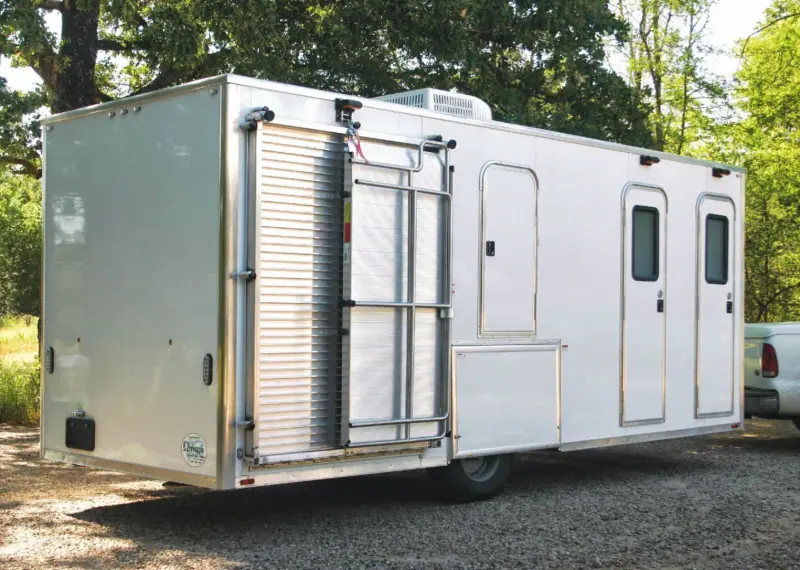 A white restroom trailer with three entry doors, metal steps, and a water tank, parked outdoors on gravel near trees.