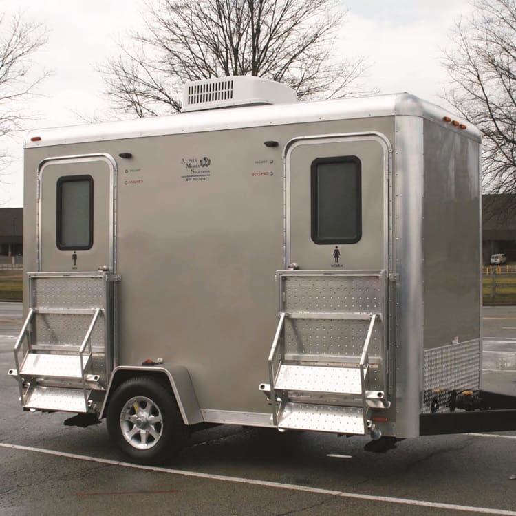 An Alpha II Restroom Trailer - A12RT with two doors and metal steps is parked in a lot with bare trees and buildings in the background.