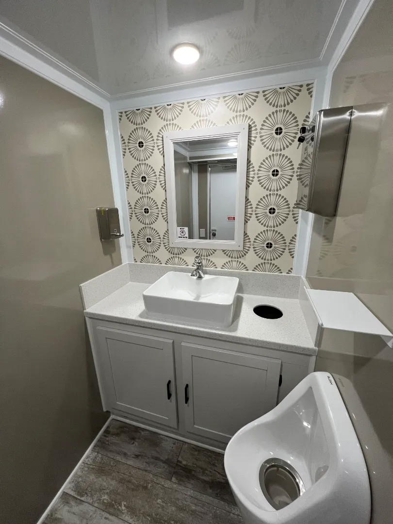Clean ada restroom trailer featuring a sink, mirror, patterned wall, urinal, paper towel dispenser, and white cabinetry on a wood-patterned floor.