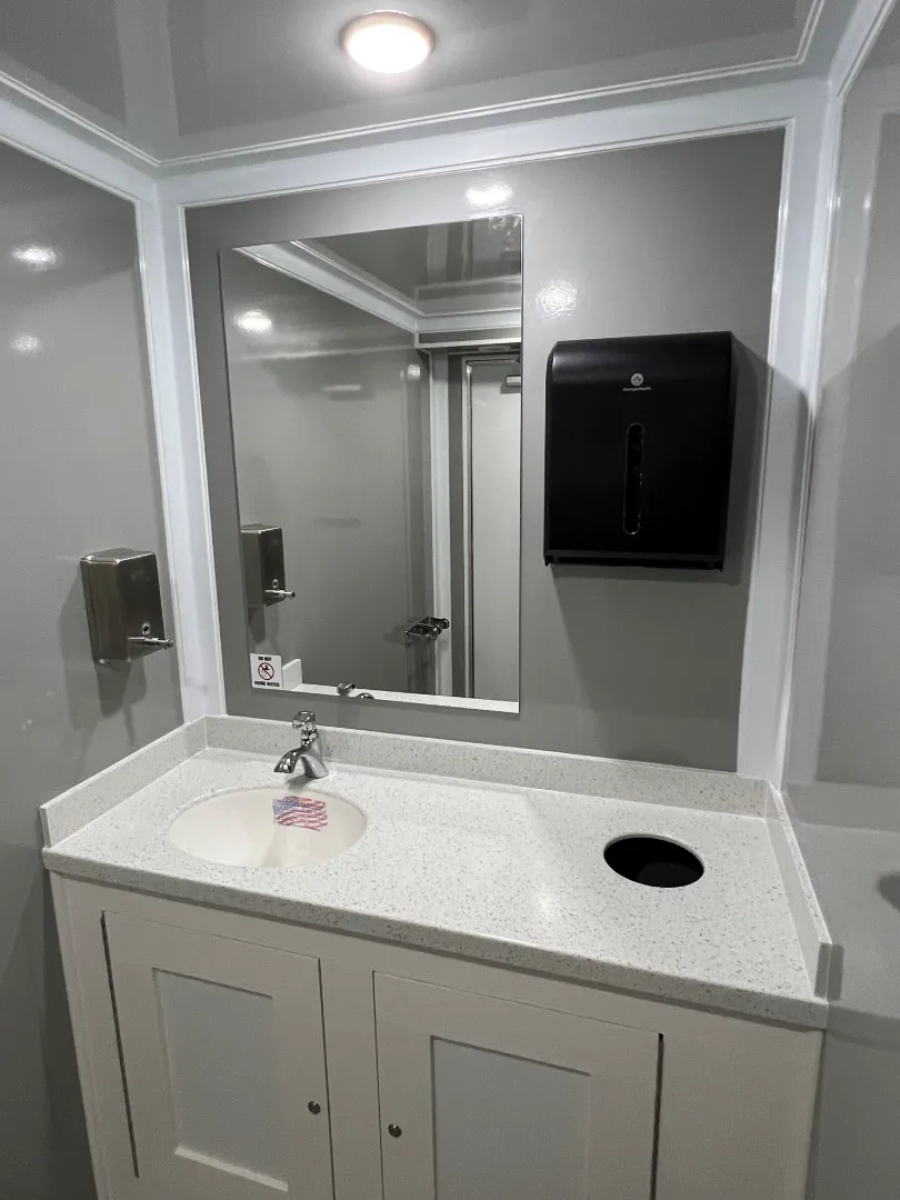 A restroom trailer features a sink area with a countertop, mirror, built-in soap dispenser, paper towel dispenser, and waste hole—designed for comfort and convenience.