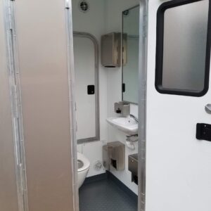 A compact ADA restroom with a toilet, sink, mirror, and mounted dispensers, viewed from the doorway. The interior of this restroom trailer features grey flooring and white walls.
